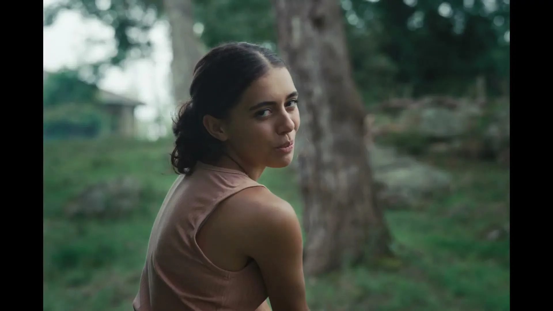 A young woman with dark hair in a bun, wearing a light brown tank top, turns her head to look back at the camera in a lush green outdoor setting.