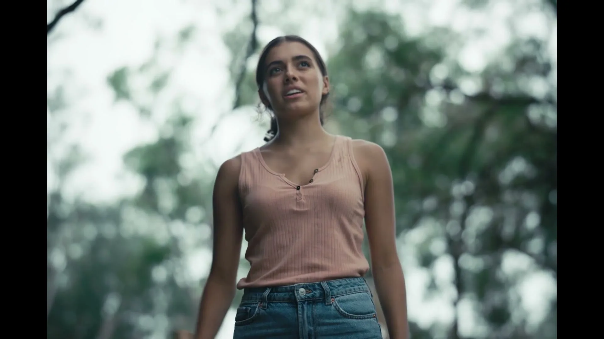 A young woman with dark hair, wearing a pink tank top and jeans, stands looking up in a forest, appearing concerned.