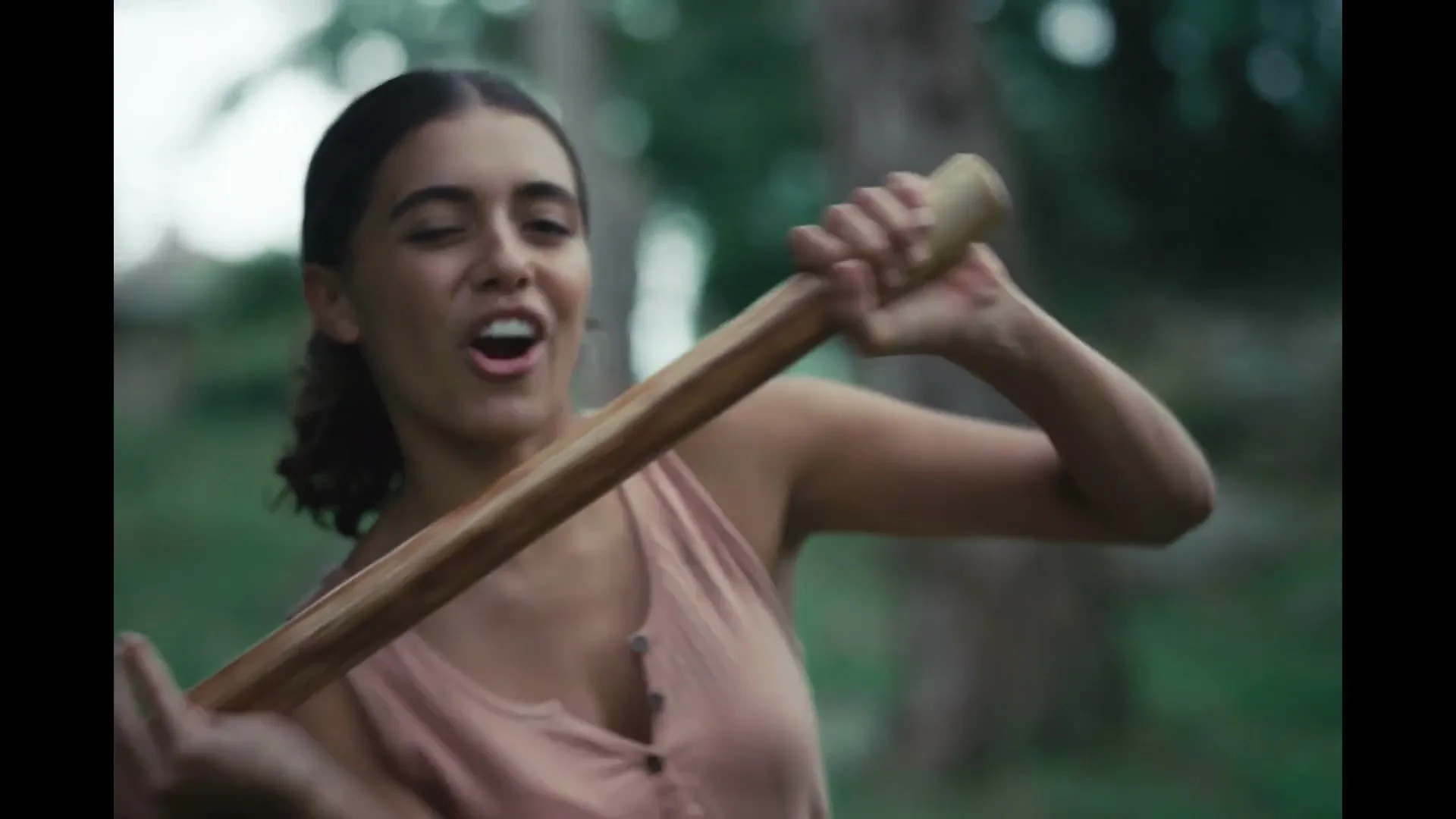 A young woman in a pink tank top swings an axe with a determined expression in a lush outdoor setting.