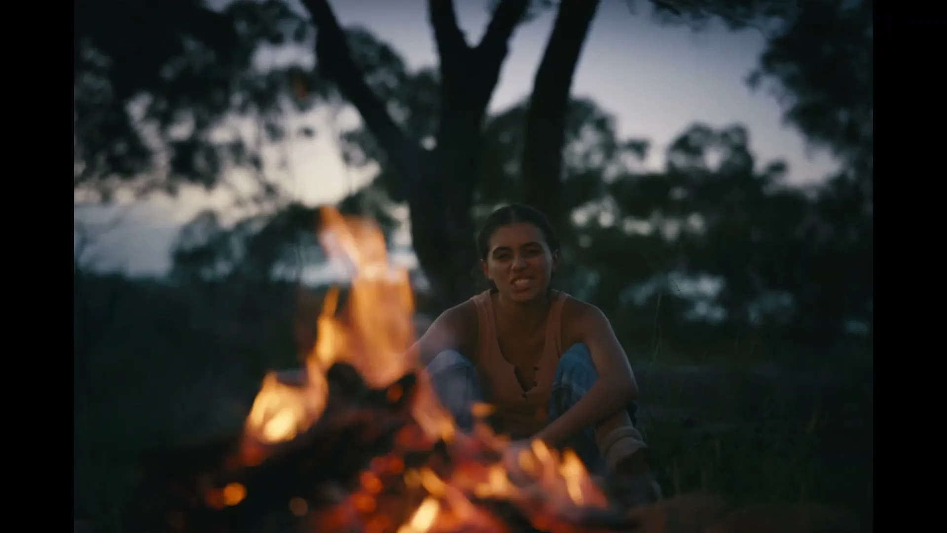 A young woman with dark hair sits by a campfire at dusk, looking directly at the camera with a serious expression.