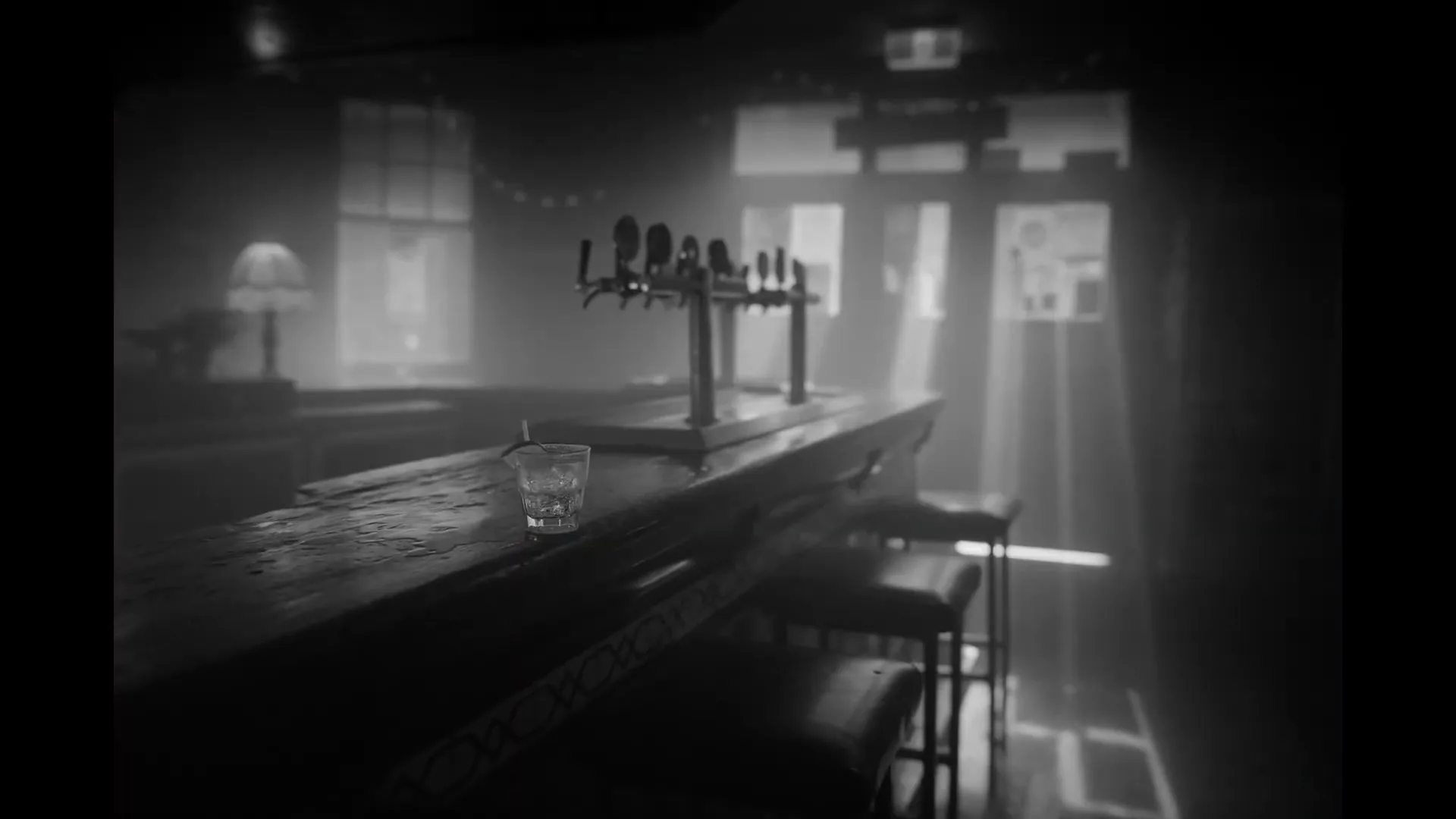 A black and white shot of an empty, dimly lit bar with a single glass on the counter, illuminated by light rays from a window.