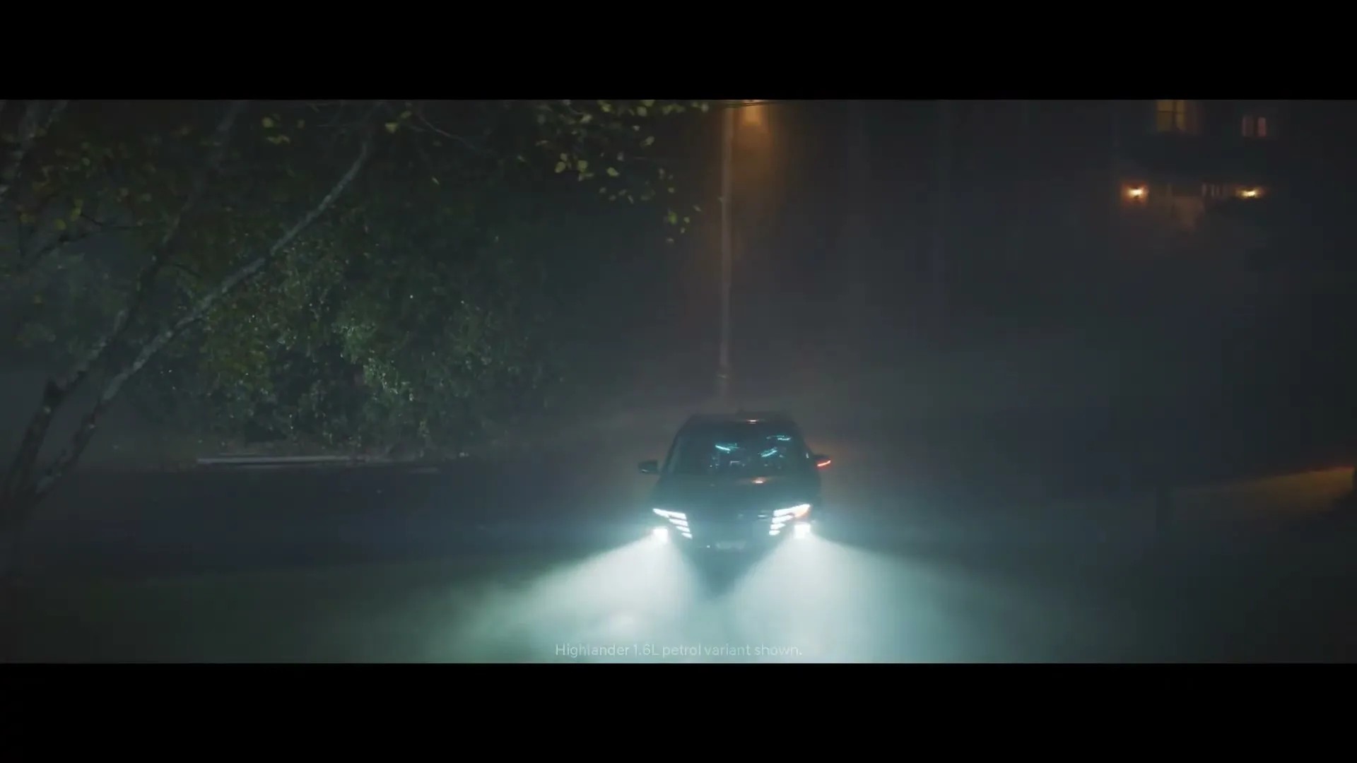 A dark Hyundai car with bright headlights drives slowly through a foggy, dark suburban street at night, with a streetlamp visible.