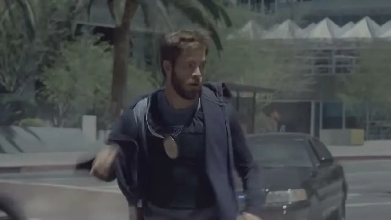 A man with a beard and dark clothing runs across a city street, with buildings and palm trees in the background.