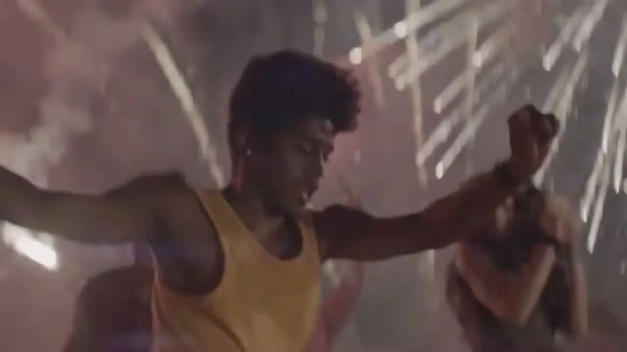 A young man in a yellow tank top dances energetically with other people in the background, surrounded by fireworks at night.