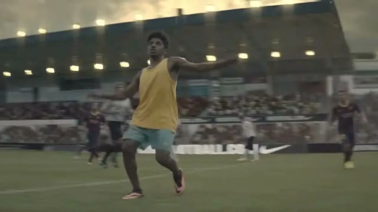 A young man in a yellow tank top and blue shorts performs a bicycle kick on a soccer field, with a stadium full of spectators in the background.