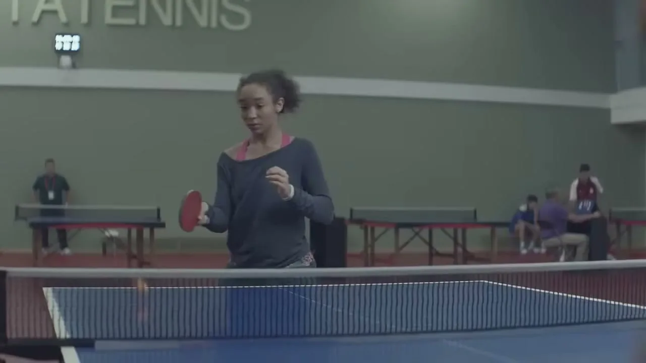 A young woman with curly hair, wearing a dark long-sleeved shirt and pink sports bra, plays table tennis in an indoor sports hall.