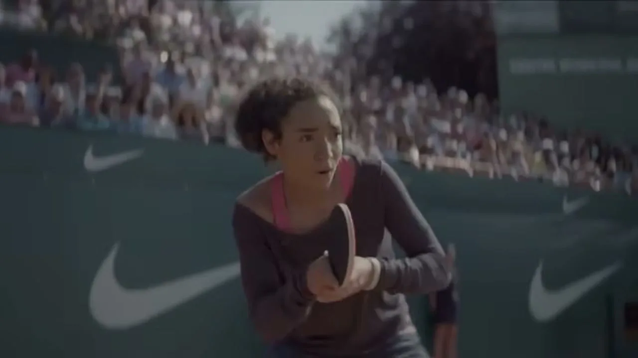 A young woman with curly hair plays tennis on a clay court, holding a paddle, with spectators in the background.