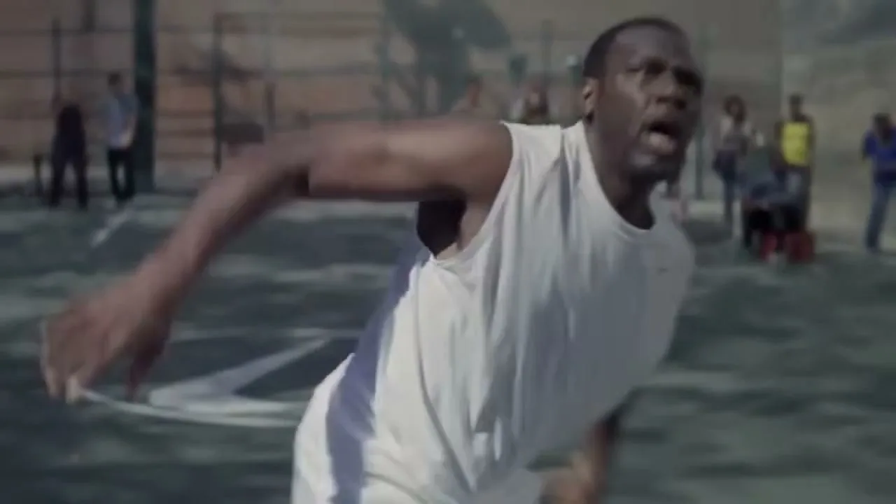 A male basketball player in a white tank top runs on an outdoor court, looking determined with other players blurred in the background.