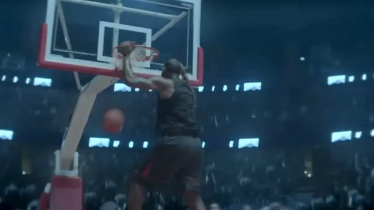 A male basketball player in a dark uniform dunks a basketball in a brightly lit stadium, with a crowd visible in the background.