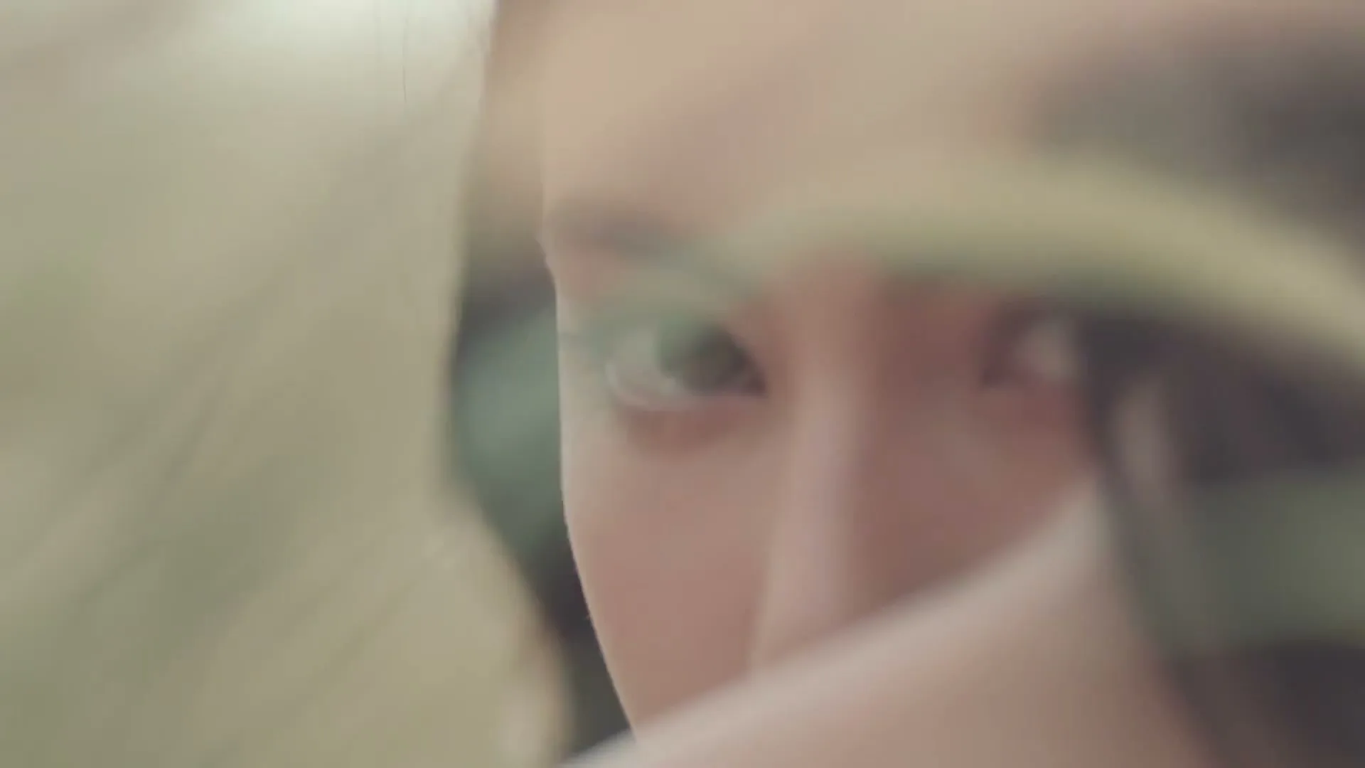 A young woman's face is partially obscured by a blurry foreground element, as she looks directly at the camera with a neutral expression.