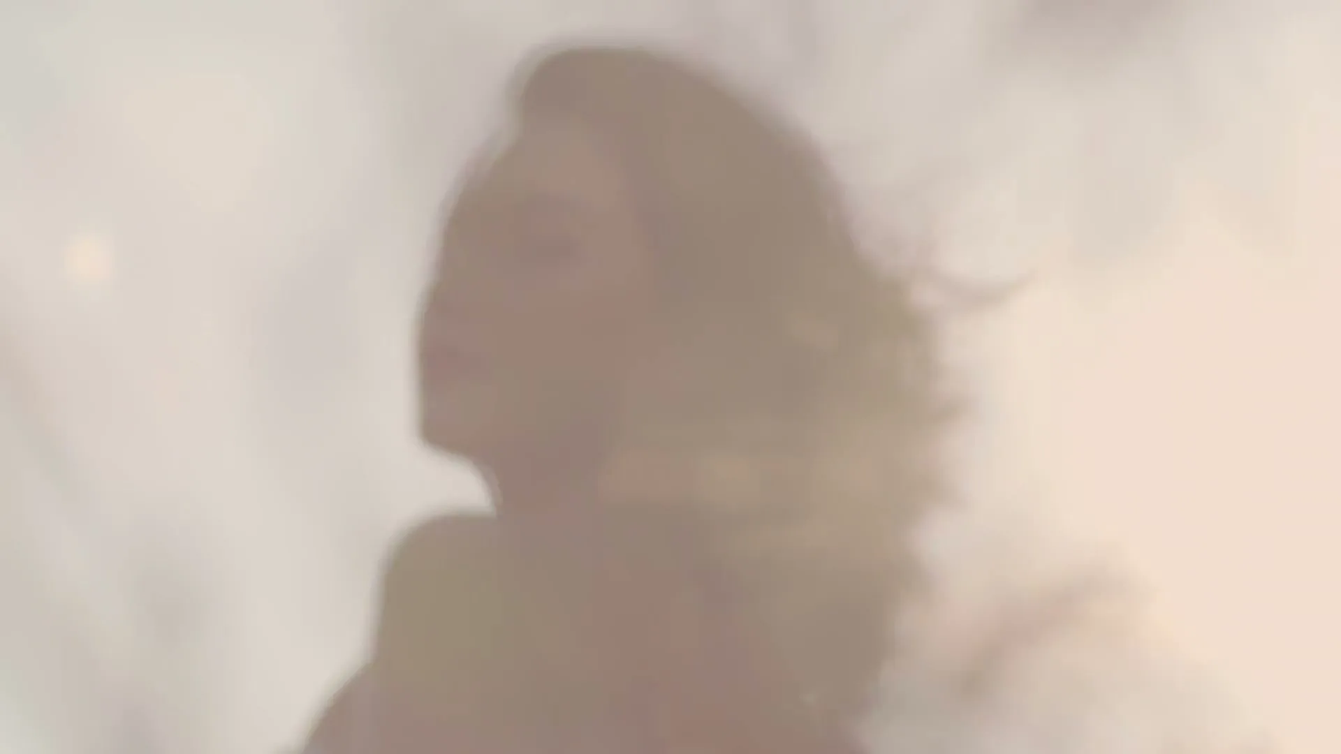 A blurry shot of a woman with dark hair, looking up and to the right, with sunlight flaring behind her, in an outdoor setting.