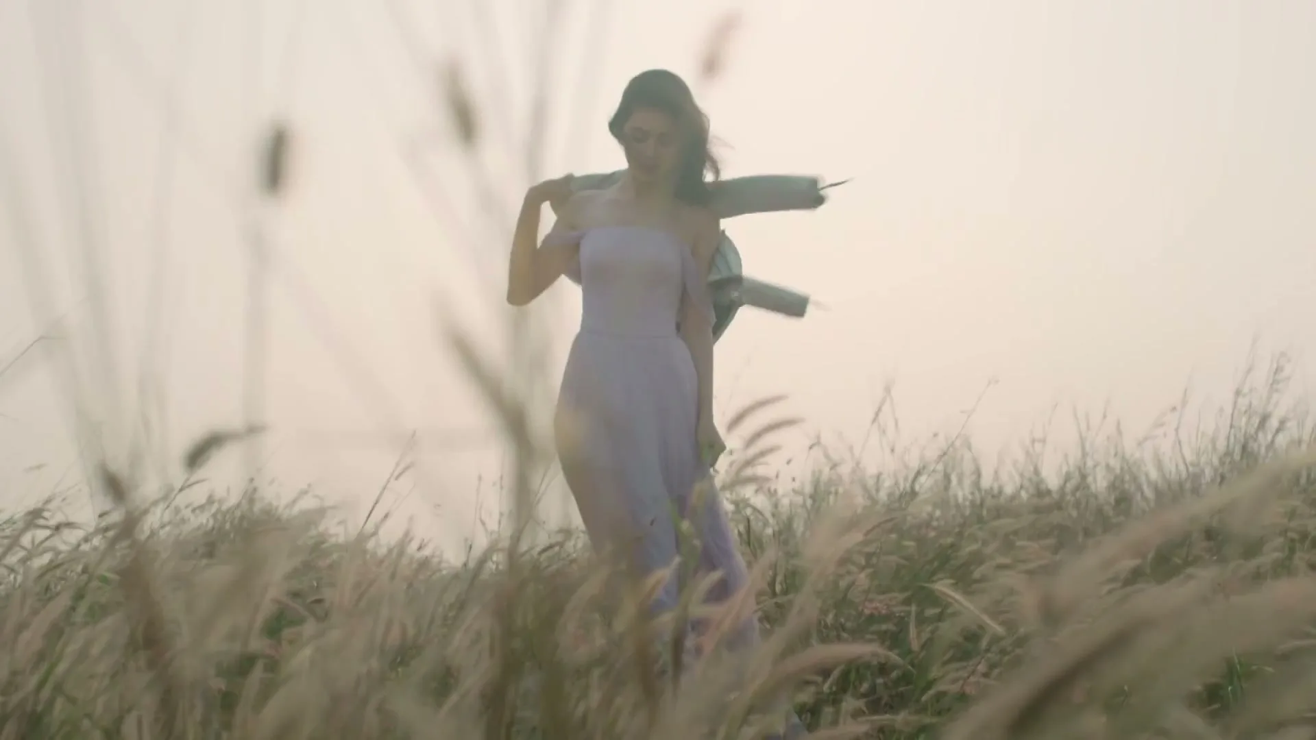 A young woman in a lavender dress and light blue jacket stands in a field of tall grass, looking down, with her hair blowing in the wind.