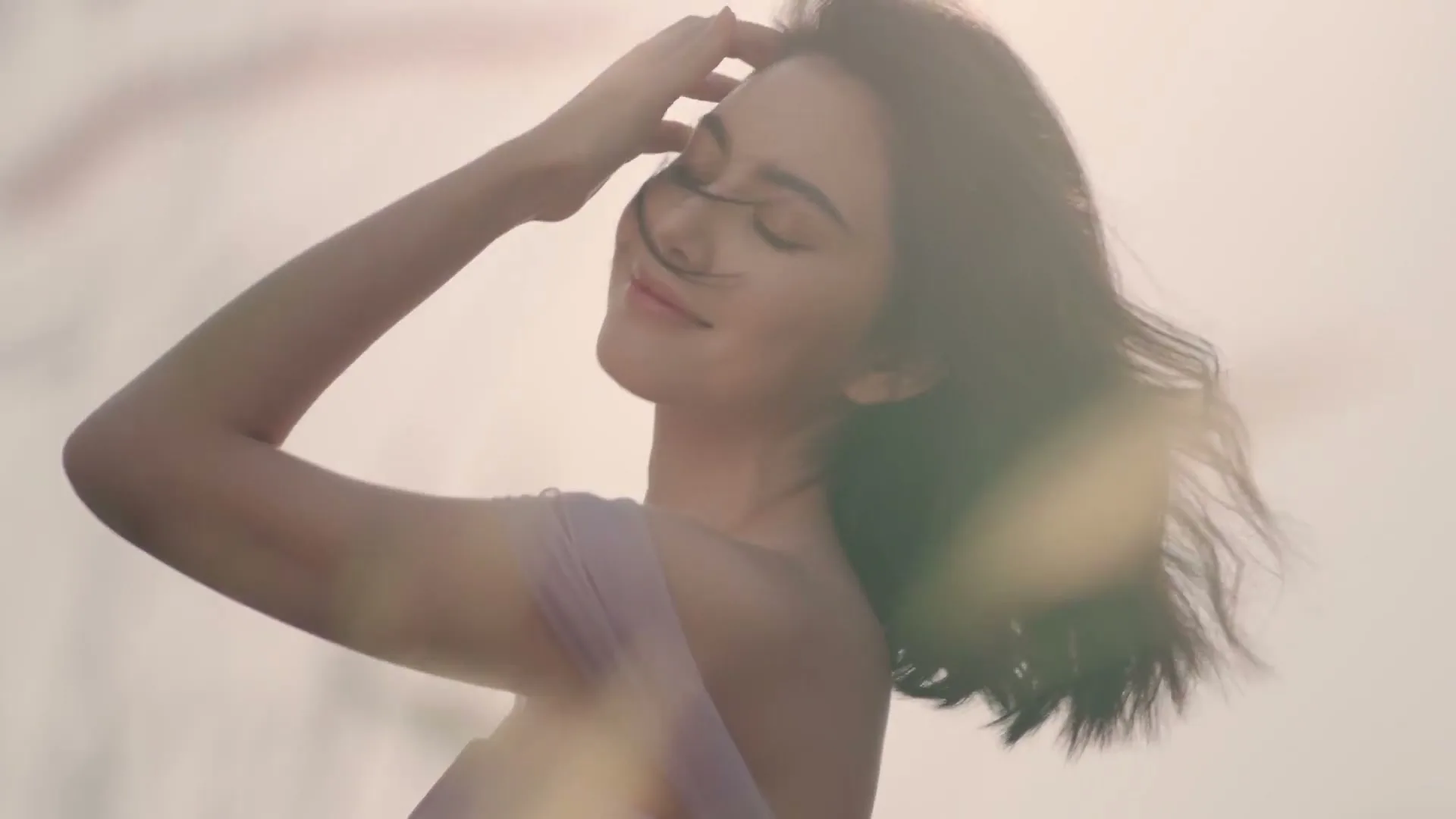 A young woman with dark hair, wearing a light purple dress, stands with her eyes closed, hand to her forehead, hair blowing in the wind, outdoors.