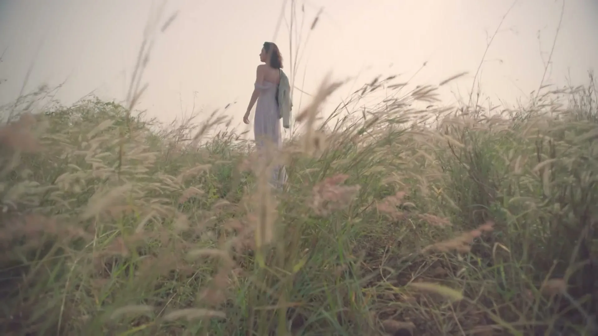 A young woman in a light dress stands in a field of tall grass, looking over her shoulder at the camera.