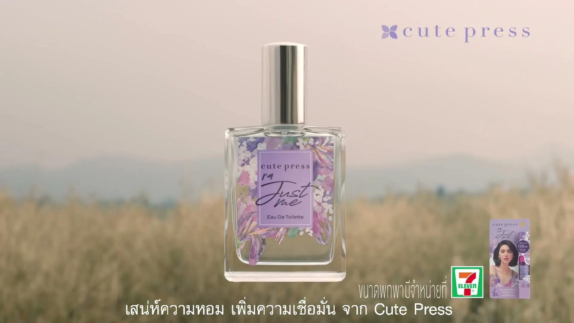 A perfume bottle with a purple floral label stands in a field of dry grass, with text and logos on screen.