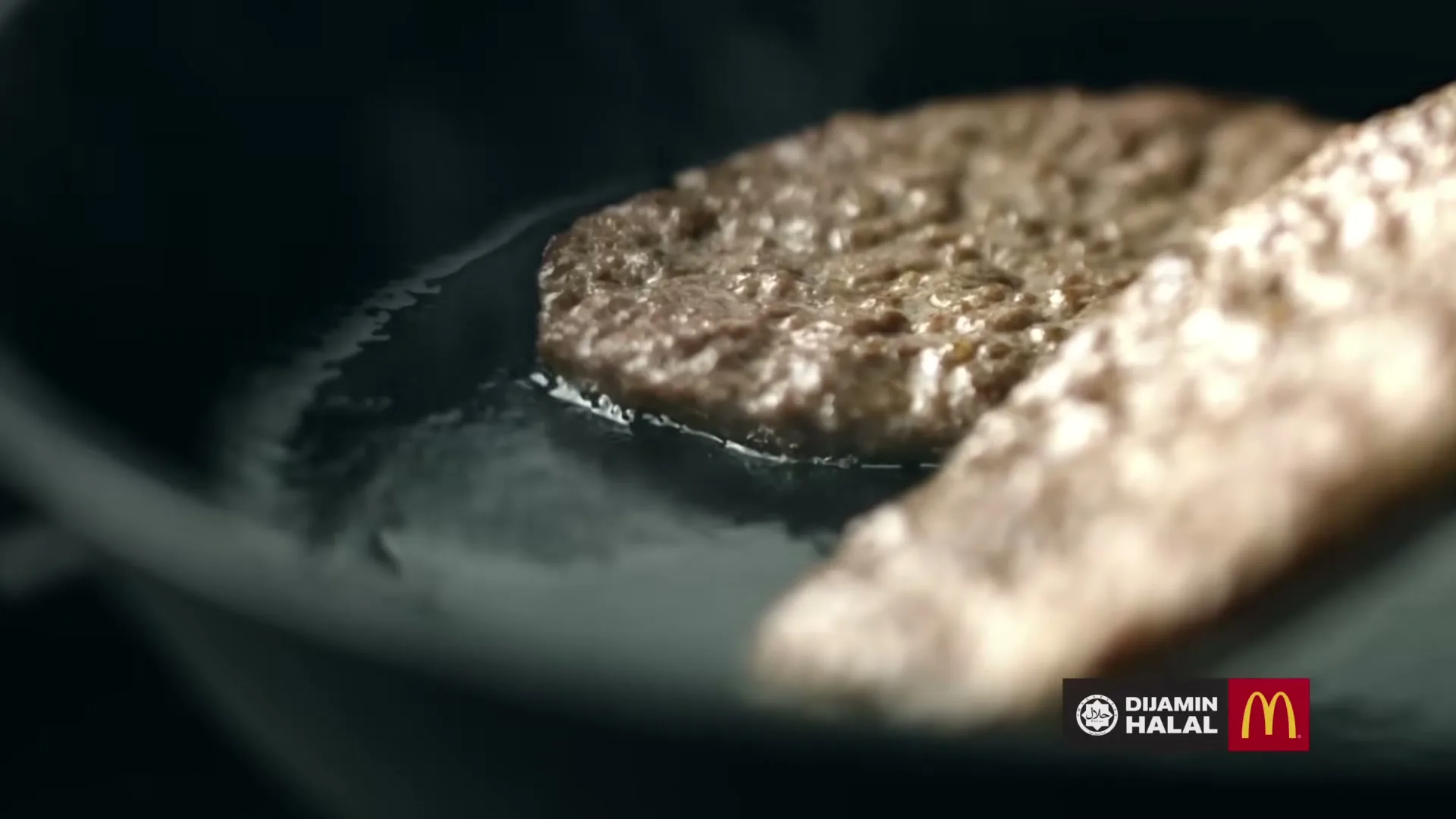 Two beef patties sizzle in a hot pan, with one patty in focus and the other slightly blurred in the foreground, in a close-up shot.