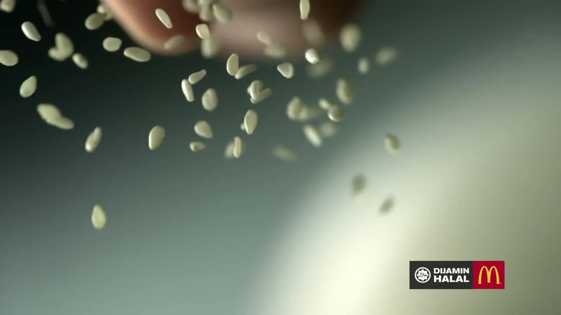A hand sprinkles sesame seeds onto a surface, with a blurred background and a logo in the bottom right corner.