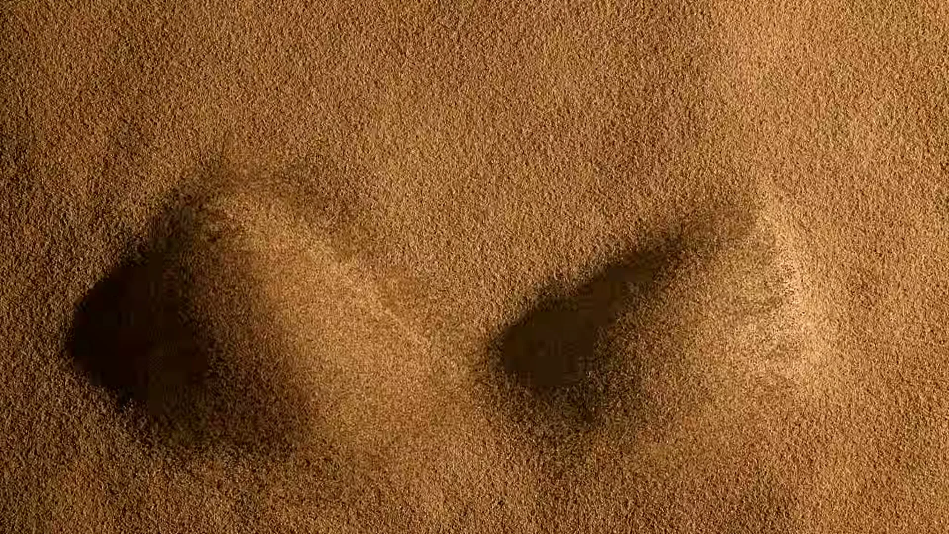 Two sand dunes emerge from a flat sandy surface, then two hands with rings and bracelets appear from the top of the frame and draw patterns on the dunes.