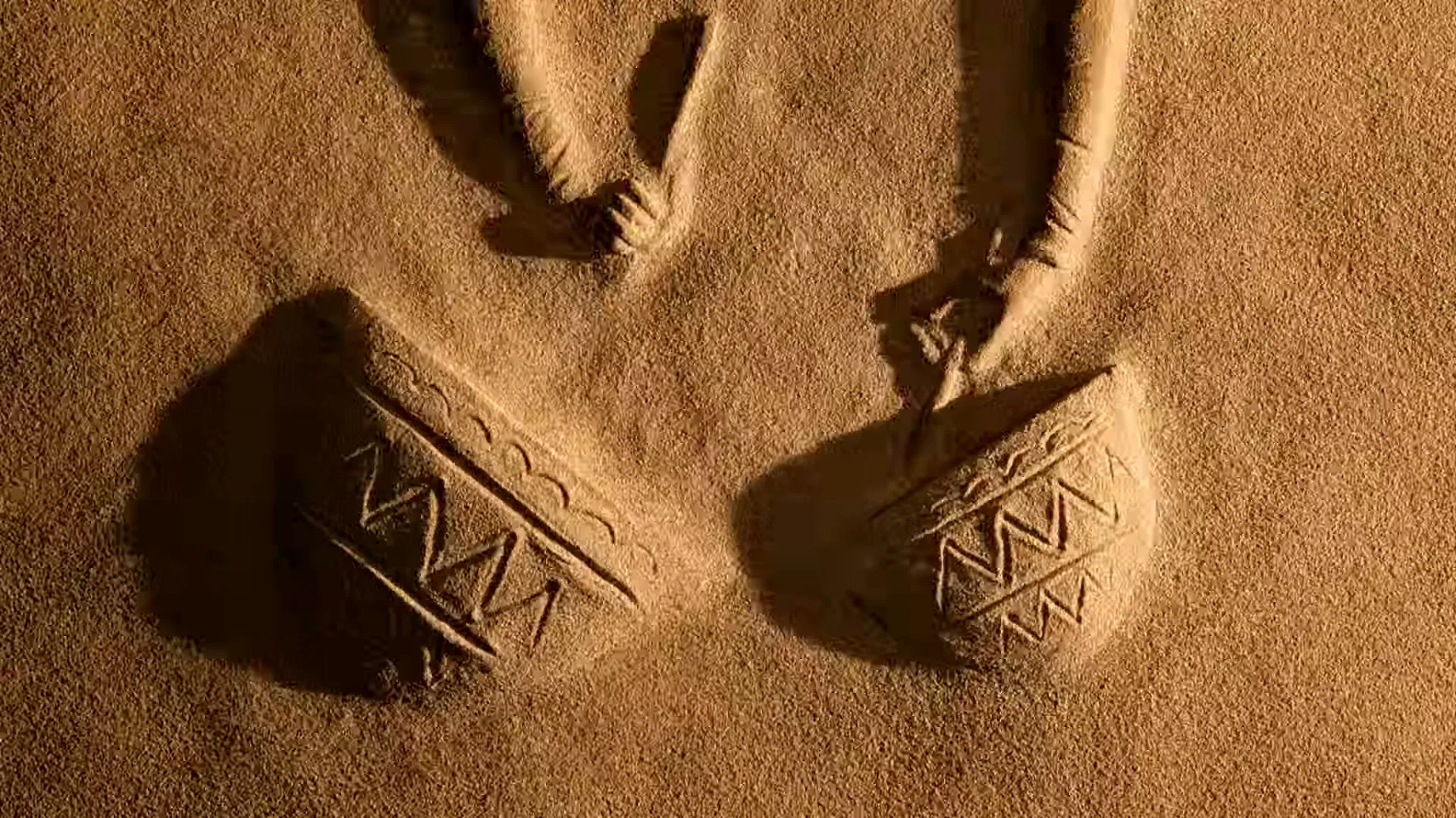 Two hands draw patterns in the sand, creating shapes that resemble traditional pottery, in a desert setting.