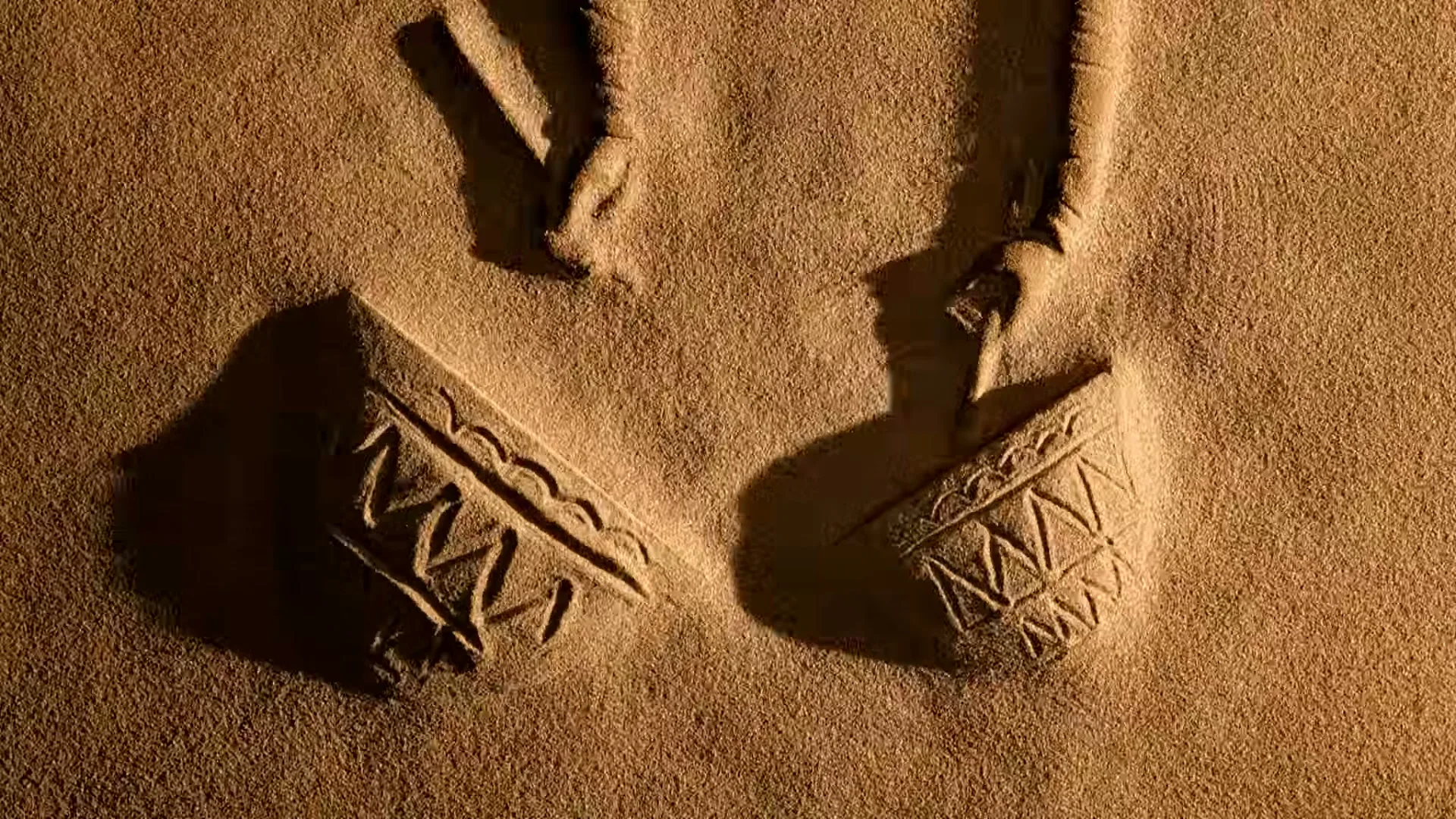 Two hands draw patterns in the sand, creating two drum-like shapes with geometric designs, in a desert setting.