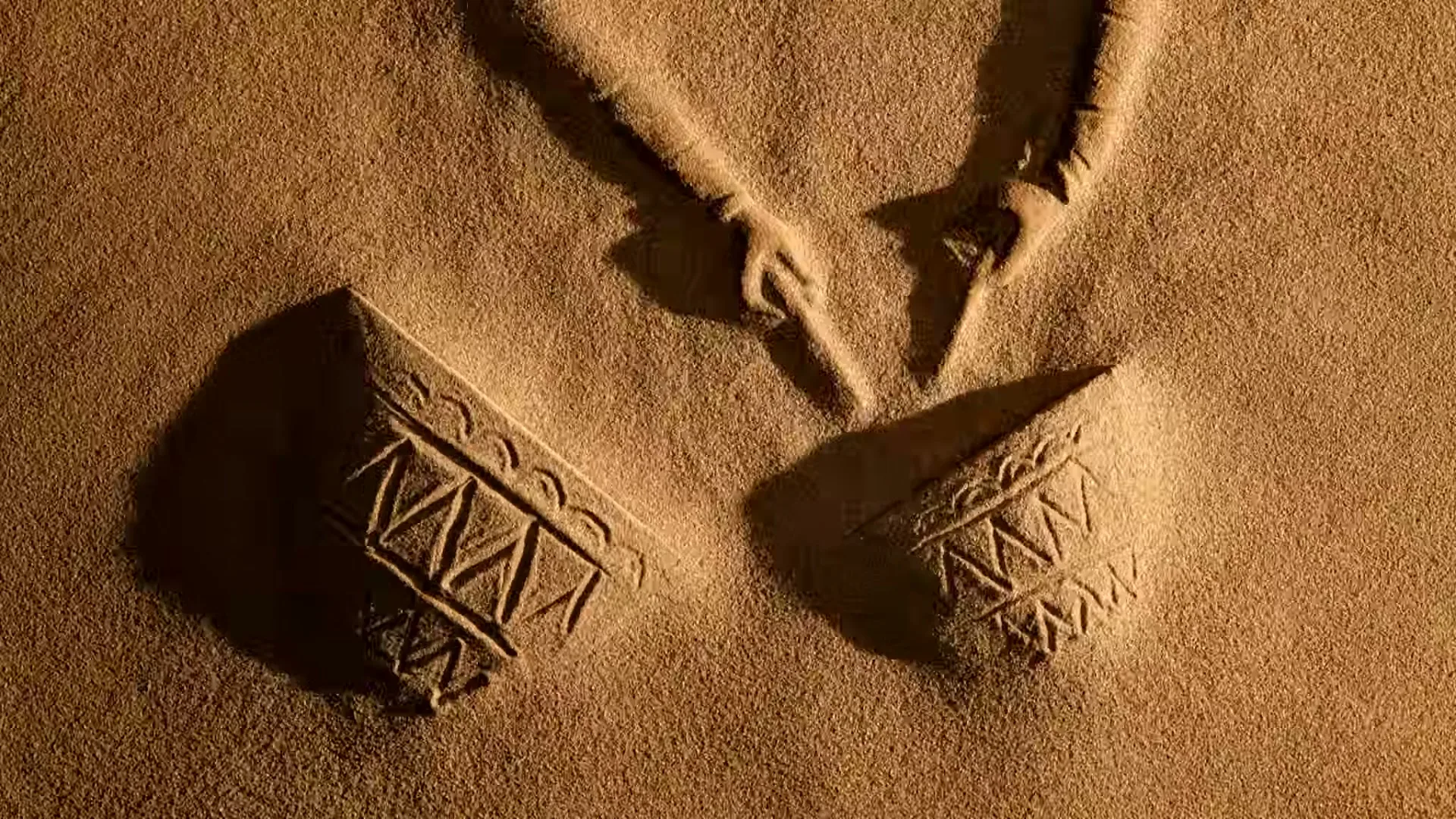 Two hands draw patterns in the sand, creating two distinct shapes that resemble traditional pottery, in a desert setting.