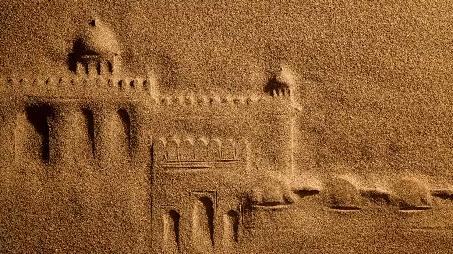 A sand drawing of a fort with camels walking past it, created by sand being blown away, in a studio.