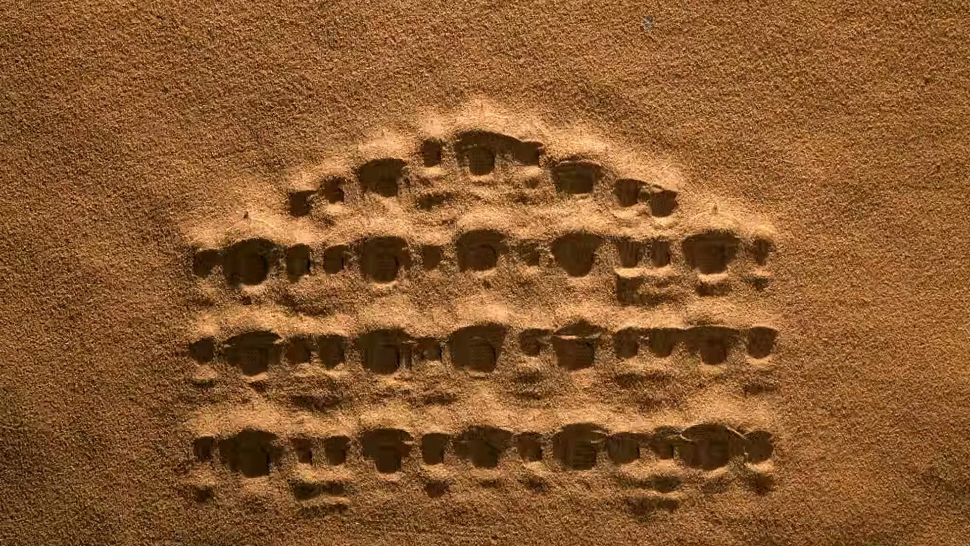 A close-up shot shows a pattern of small indentations in the sand, resembling a miniature city or fort, with sand blowing over it.