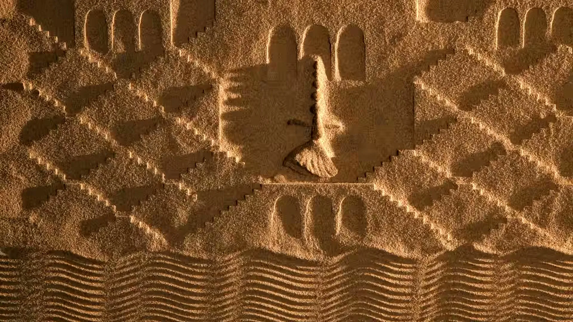 A hand draws patterns in the sand, creating a stylized representation of a stepwell and water, under warm sunlight.