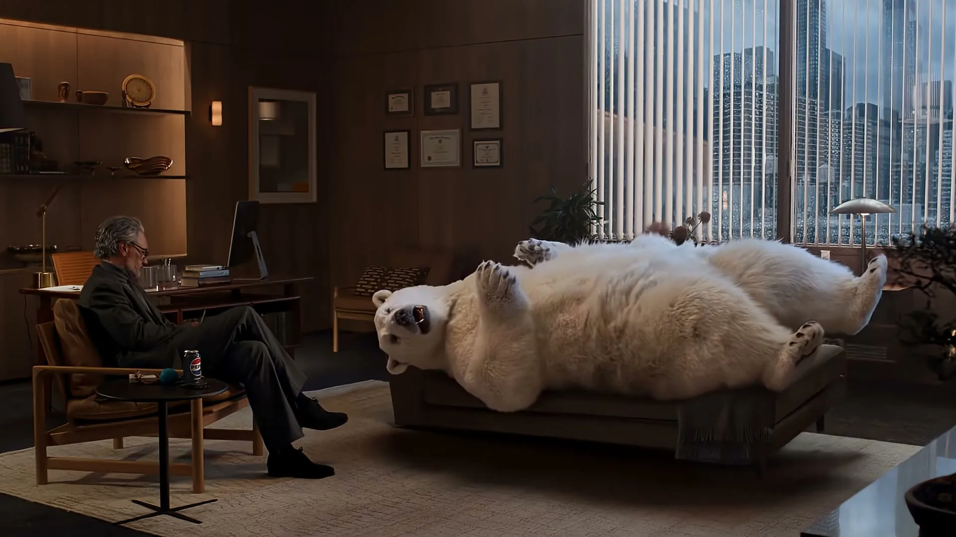 A man in a suit sits in a chair, looking at a polar bear lying on a couch in a modern office with a city view.