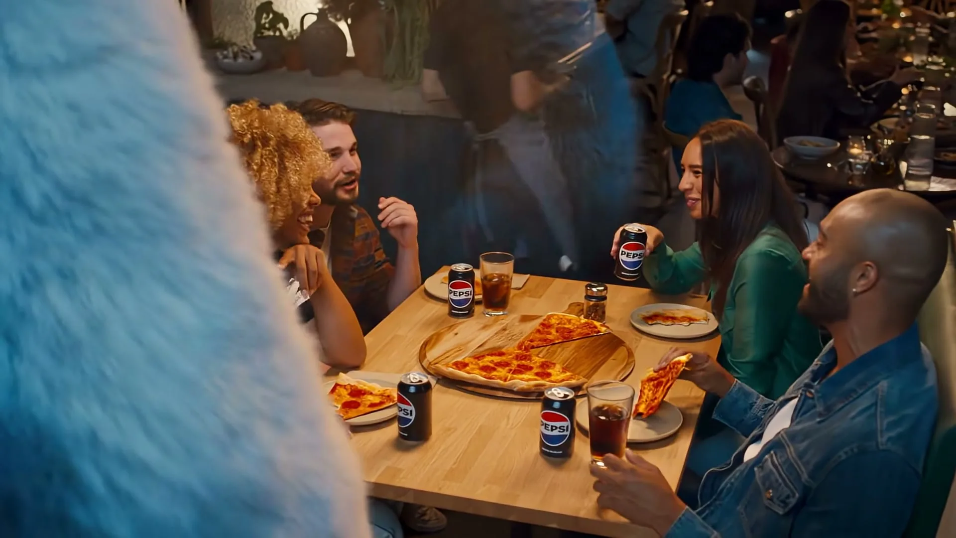 A diverse group of four friends enjoys pizza and Pepsi at a lively restaurant, with one woman taking a big sip from her can.