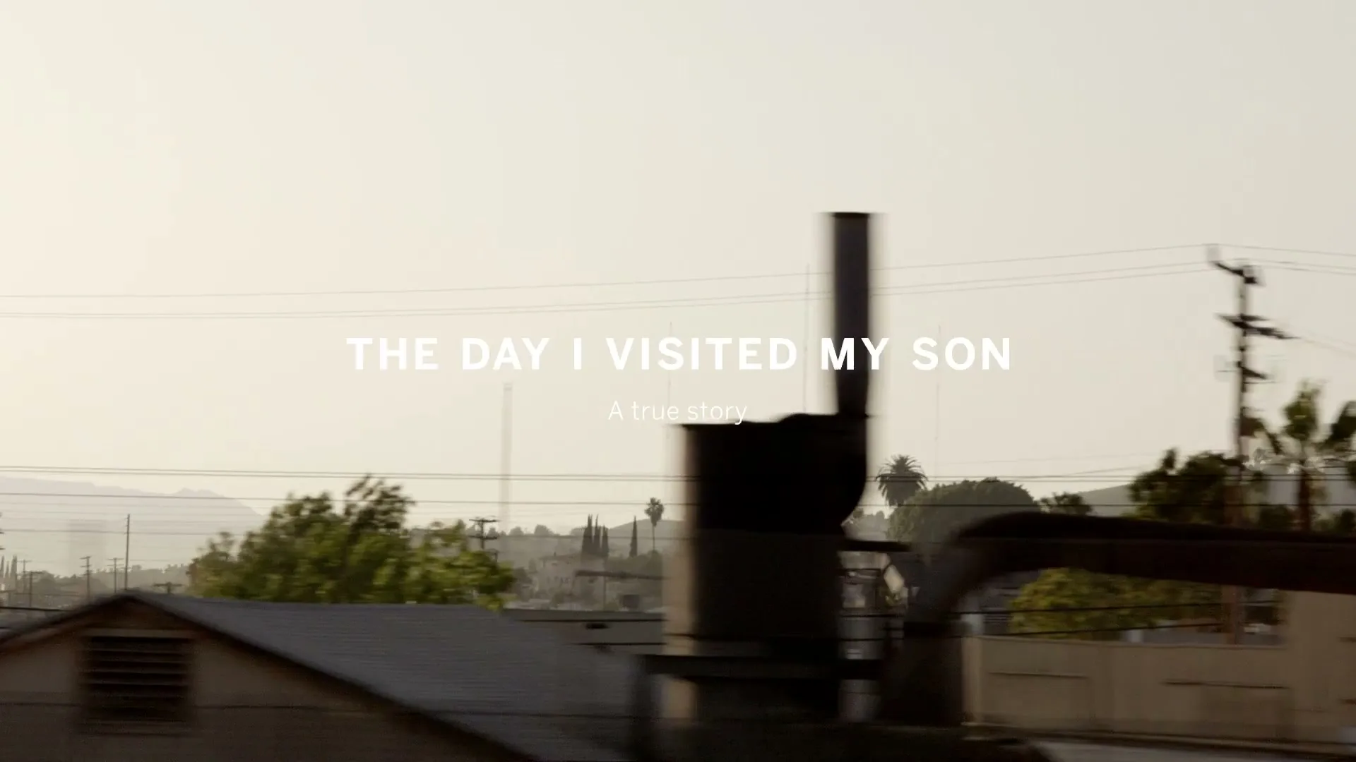 A title card with white text "THE DAY I VISITED MY SON" and "A true story" appears over a moving shot of an industrial area with buildings, power lines, and distant hills.