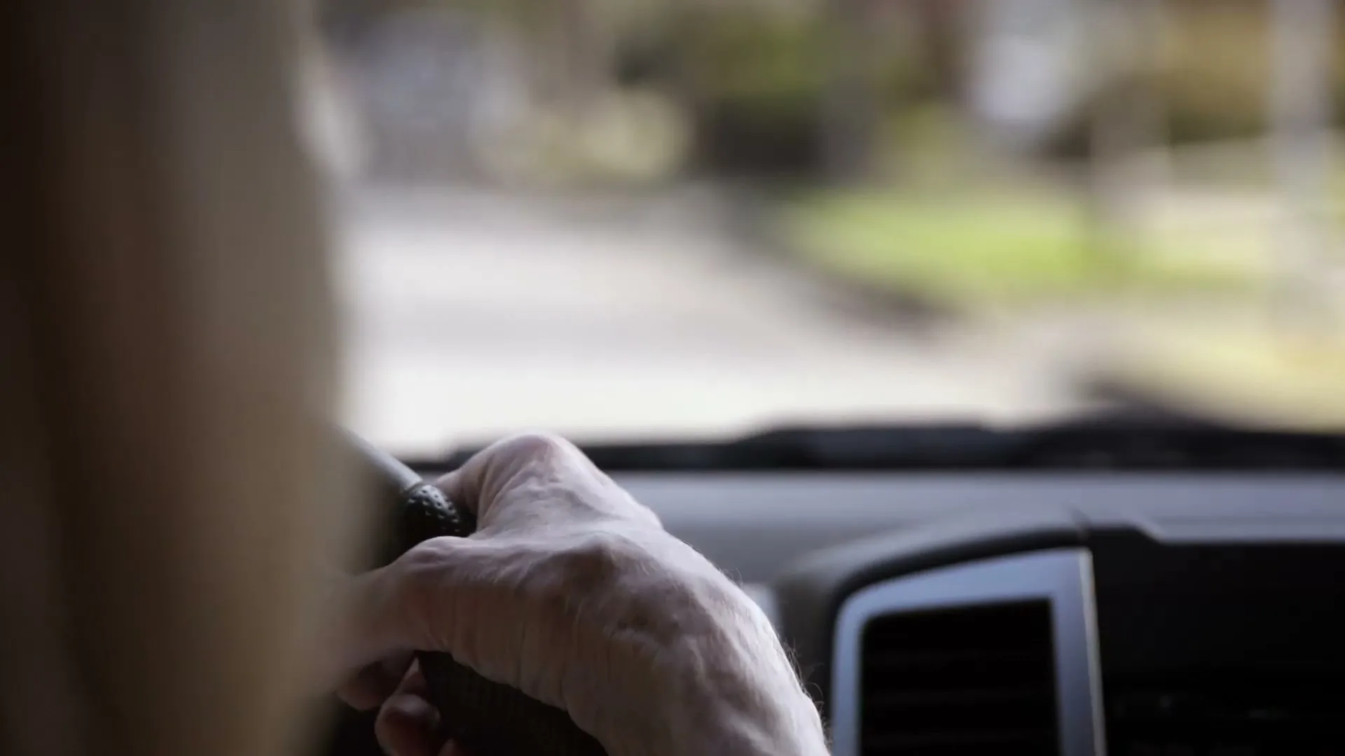 An older person's hand is seen on a steering wheel, driving a car down a blurred street during the day.