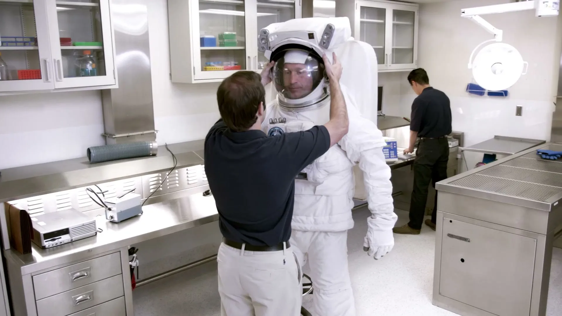 A man in a dark polo shirt helps another man in a white spacesuit put on his helmet in a sterile, white lab with metal counters.