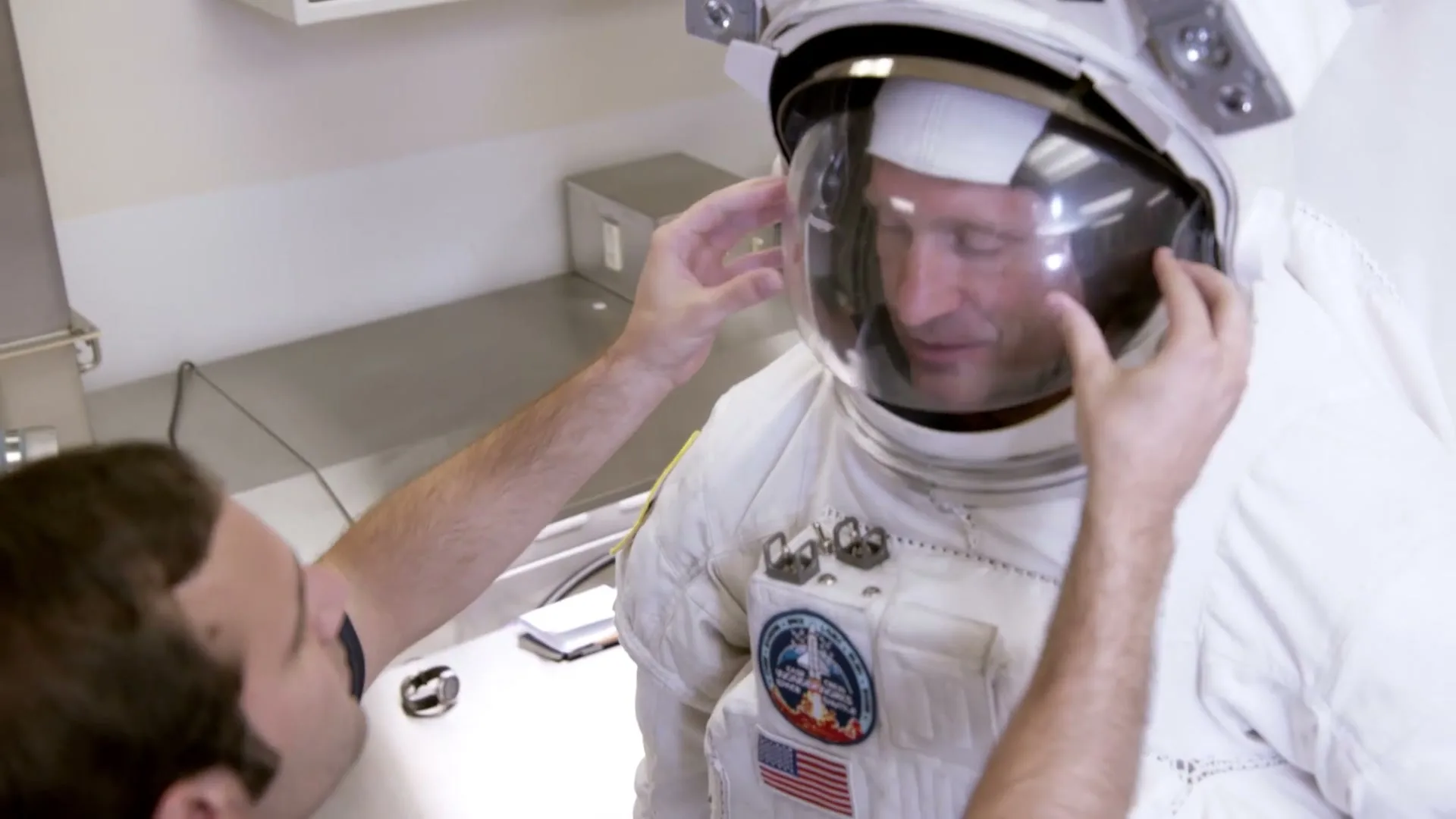 A man in a white astronaut suit has his helmet visor adjusted by another man in a brightly lit room.