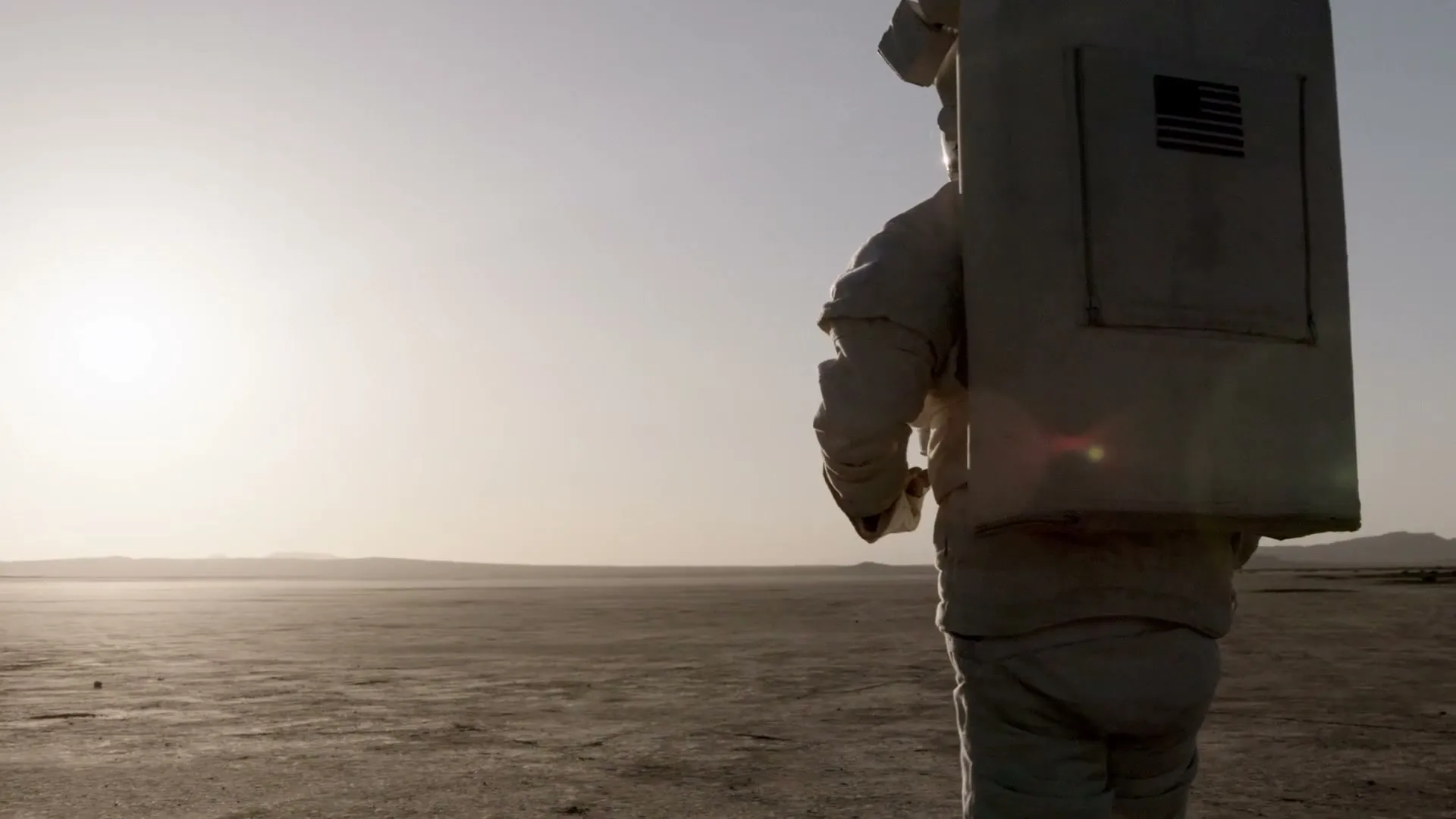 A person in a white astronaut suit with an American flag on their backpack walks across a vast, barren desert landscape under a bright sky.