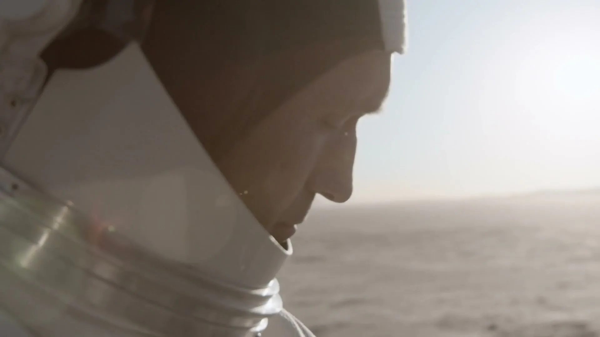 A male astronaut in a white spacesuit looks down with a pensive expression on a desolate, sandy planet under a bright sky.