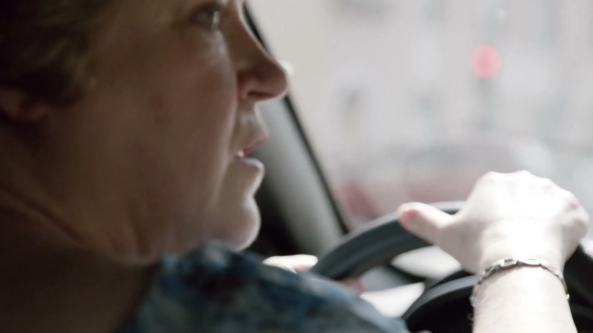 A middle-aged woman with short blonde hair drives a car, looking intently forward with her hands on the steering wheel.