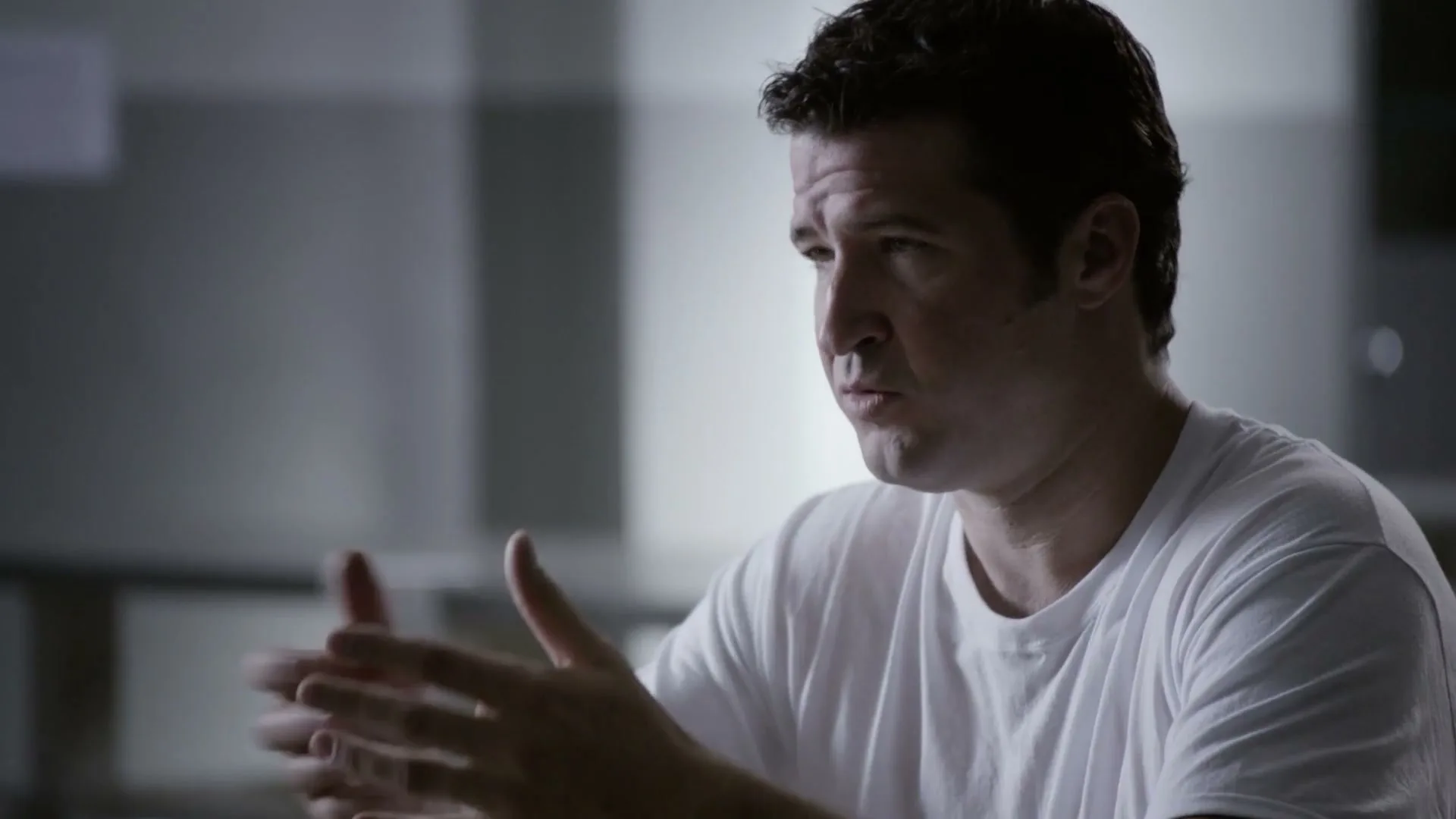 A man in his 30s, wearing a white t-shirt, sits and talks, gesturing with his hands in a dimly lit room.