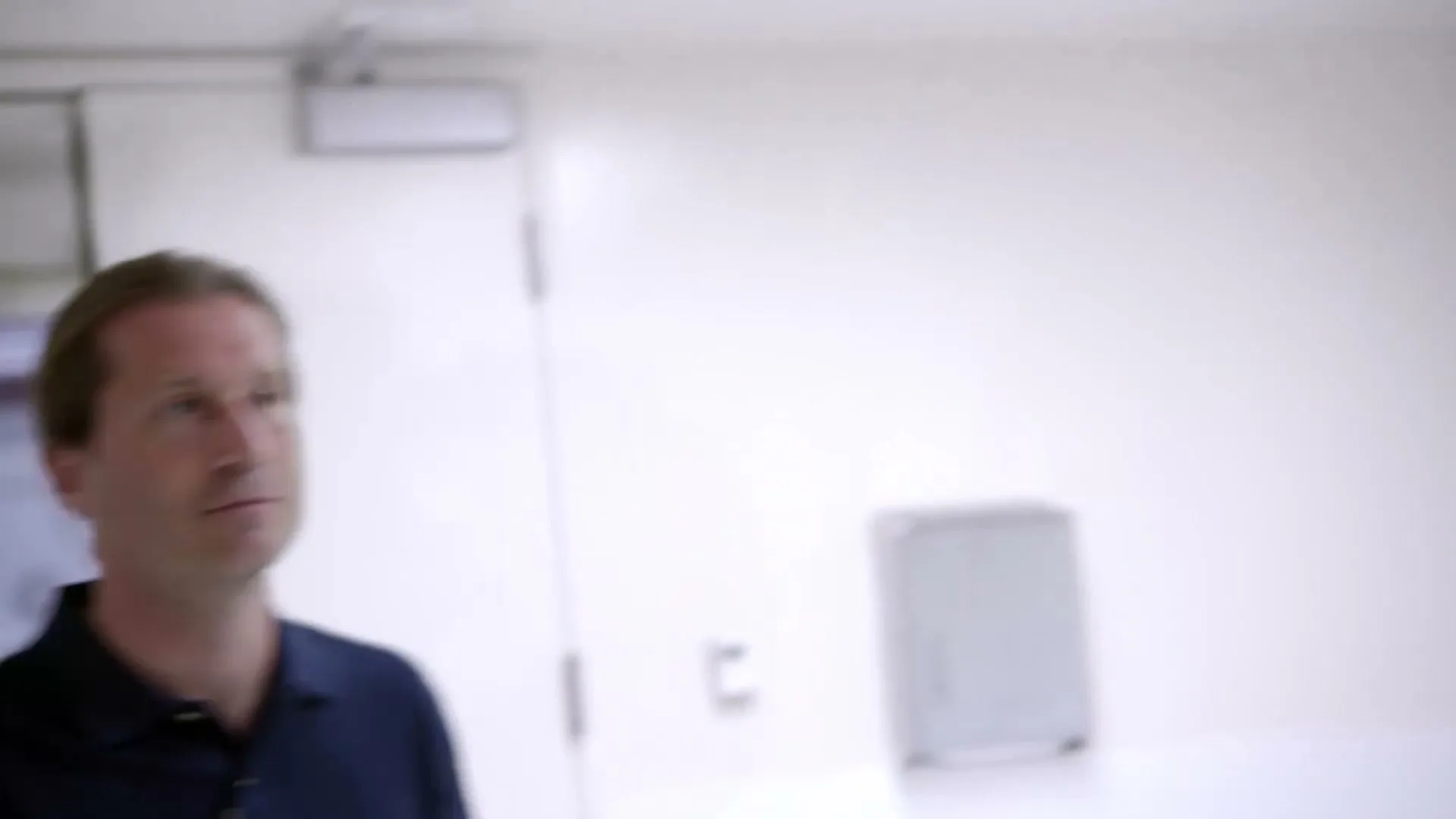 A man with long hair in a dark polo shirt walks through a brightly lit, white hallway, looking up and to the right.