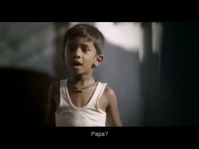 A young boy, wearing a white undershirt and a necklace, looks up with a worried expression in a dimly lit interior.
