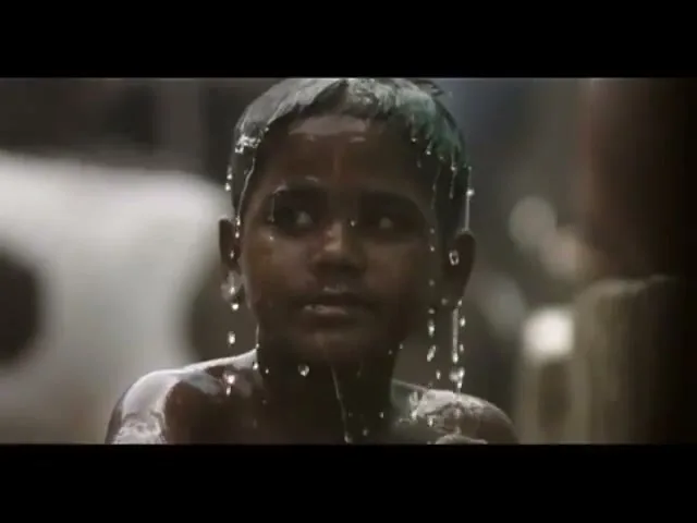 A young child, with dark skin and wet hair, looks off-camera as water drips from their face and hair in an outdoor setting.