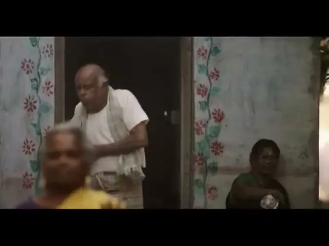 An elderly man in a white shirt and patterned sarong exits a doorway, looking concerned, while two elderly women sit nearby in a rural setting.