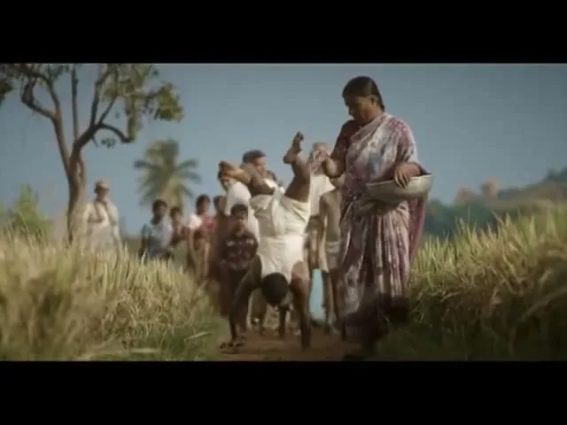 A woman in a sari walks alongside a man doing a handstand, followed by a group of people, on a dirt path through a field.