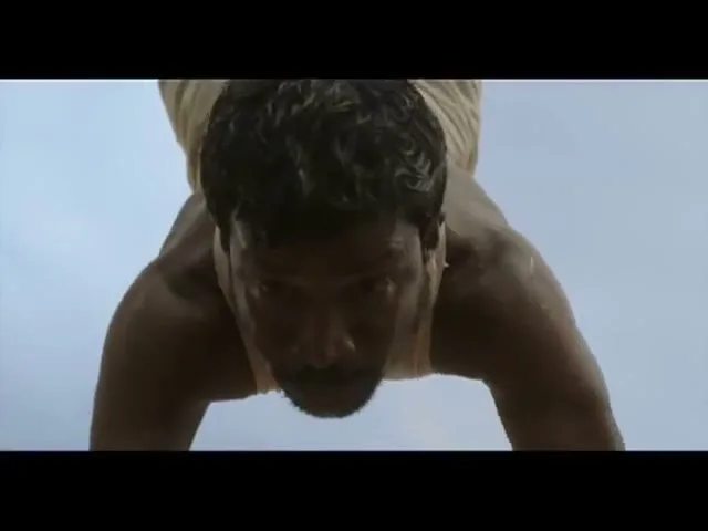 A man with dark, wet hair and a mustache is seen from below, doing push-ups against a bright, overcast sky.