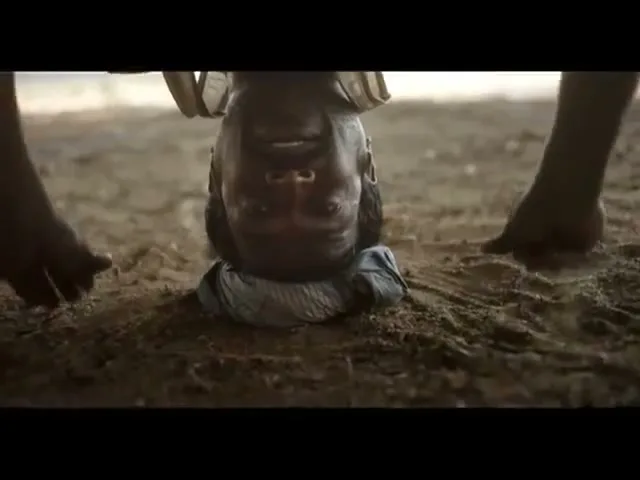 A man with dark skin and a beard is upside down, balancing on his head on a piece of cloth on the ground, with his hands and feet visible.
