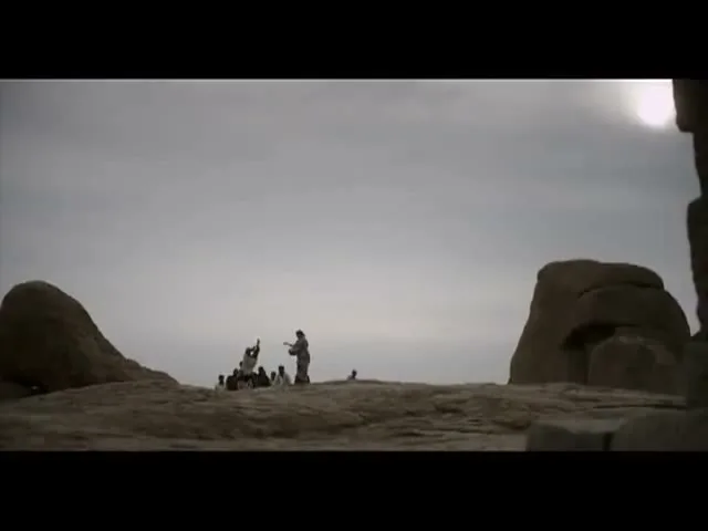 A group of people, including children, are gathered on a rocky outcrop under a vast, cloudy sky, with one person raising their arms.