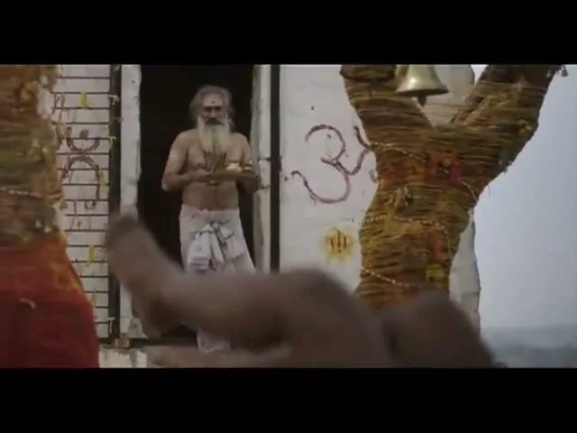 A holy man with a long white beard and a white loincloth stands in a doorway, holding a small bowl, as a hand rings a bell in the foreground.