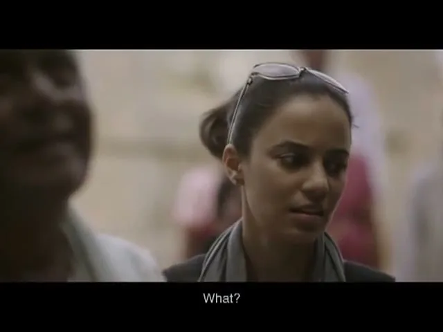 A young woman with sunglasses on her head looks confused while talking to an older man in a crowded, bright interior setting.
