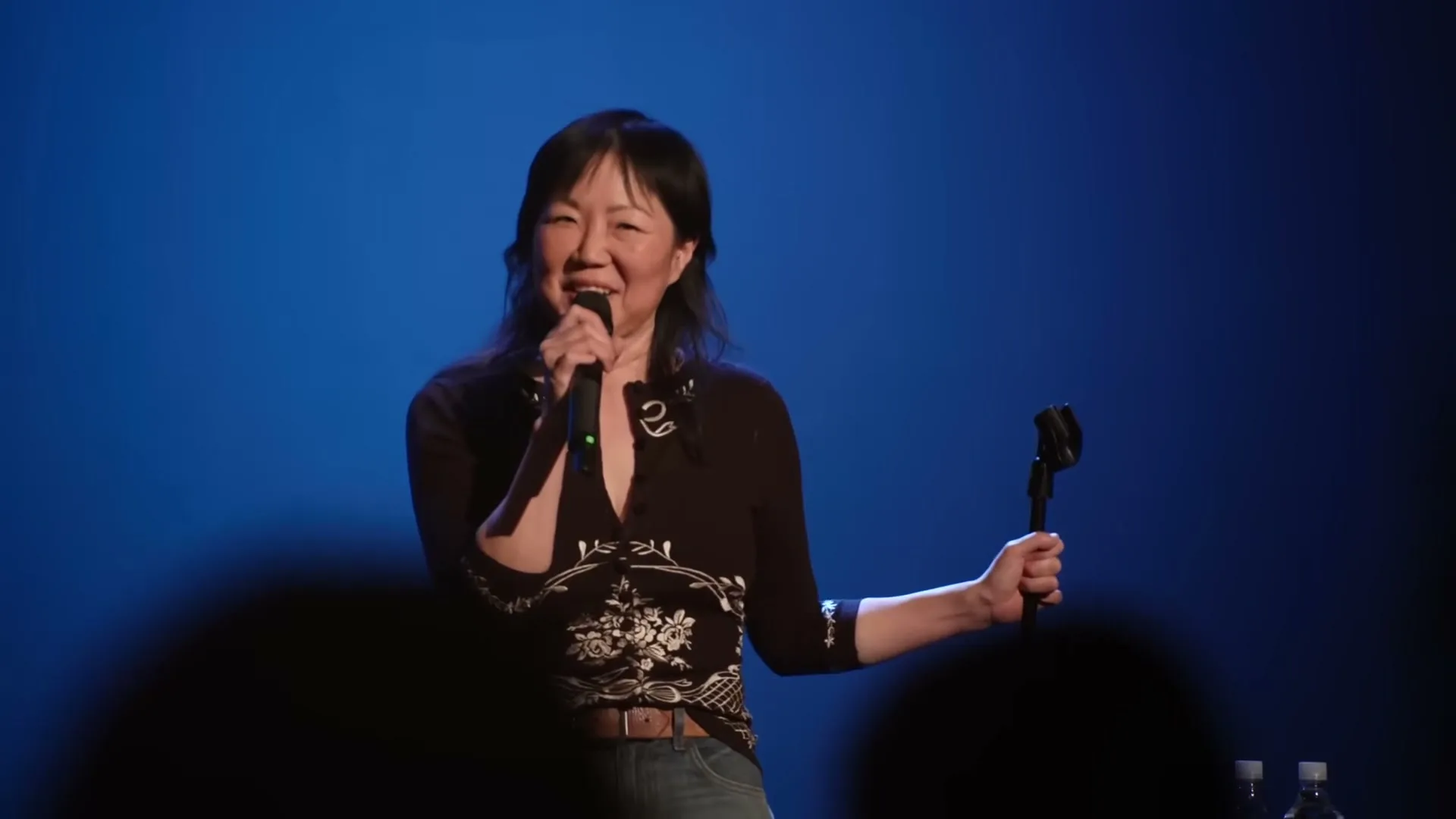 A female comedian with dark hair and a patterned shirt performs stand-up comedy on stage, holding a microphone against a blue background.