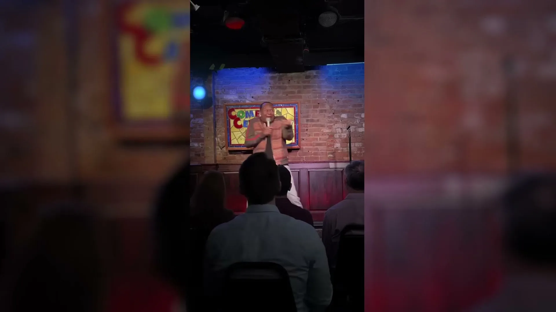 A male comedian in a patterned shirt performs stand-up comedy on stage in a dimly lit club, gesturing with his hands to an unseen audience.